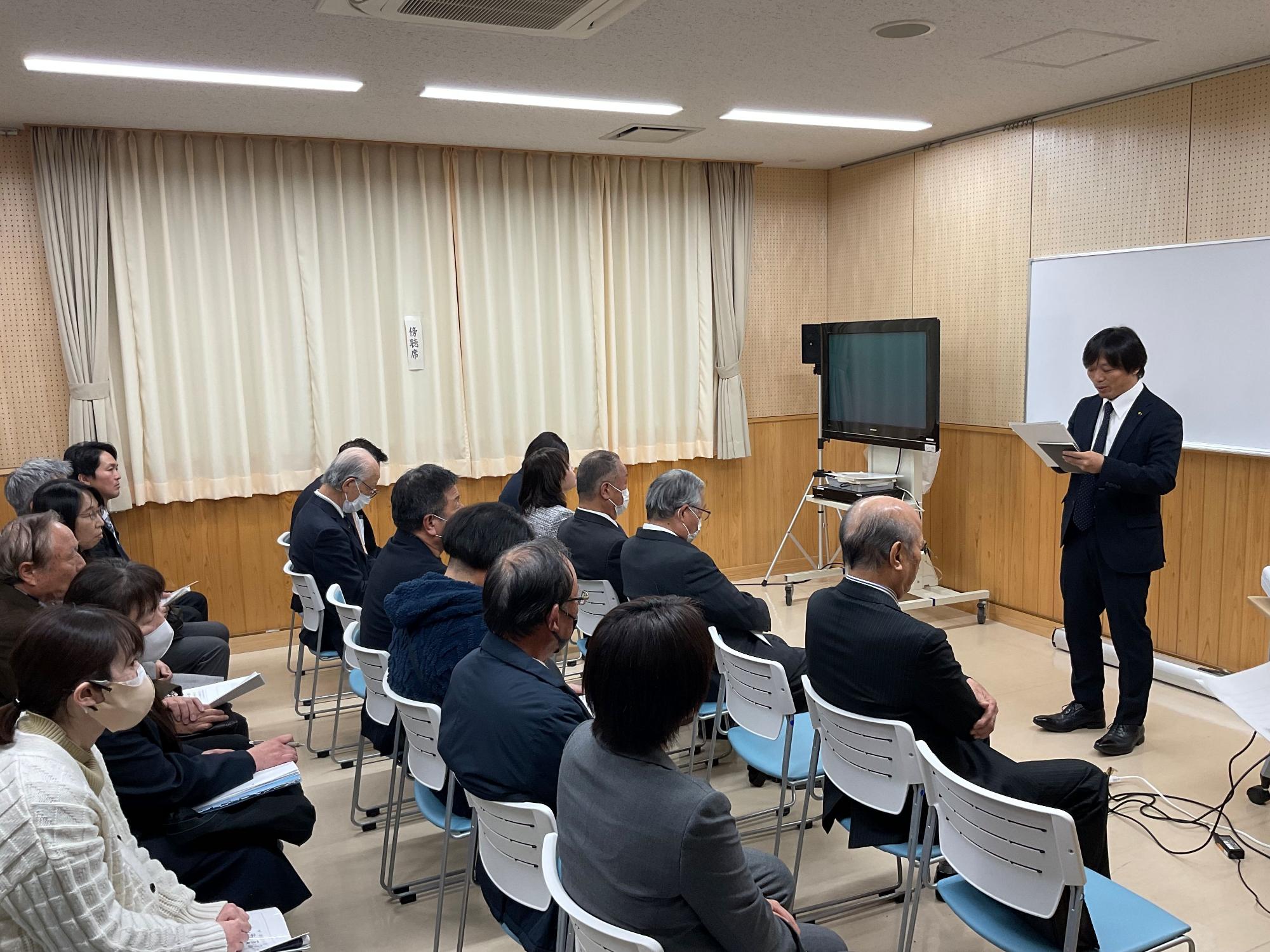 会議室のような部屋で、スーツ姿の男性が前方に立って書類を読み上げ、椅子に座った多くの聴衆が注目しており、カーテンには「式次第」の貼り紙が見える文教福祉常任委員会の様子の写真