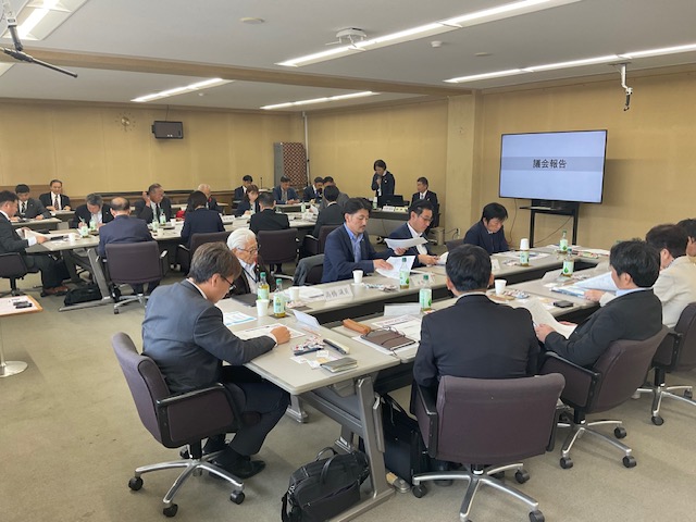 会議室で、スーツ姿の多くの人々が「議会報告」と映るスクリーン前の長いテーブルに着席し、手元の資料に目を通している真剣な会議の様子の写真