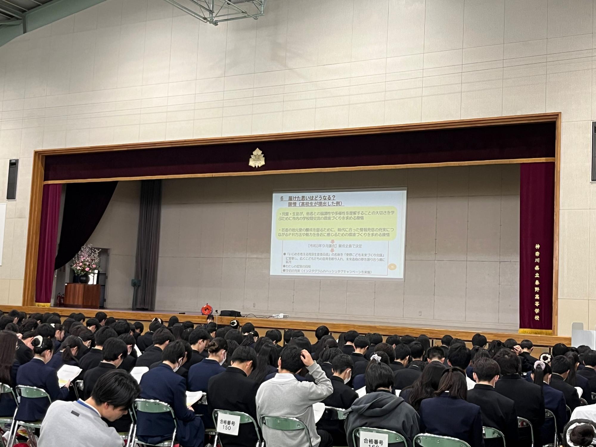 体育館のような広い会場で、大勢の生徒が黒い制服姿で座って壇上のスクリーンを見ており、スクリーンには「6 届けたい想いをカタチに」から始まる日本語のプレゼンテーション資料が投影されている出前講座の様子写真