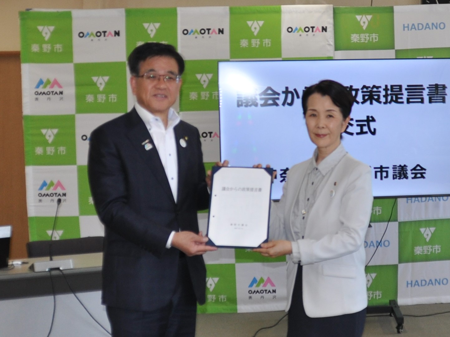 「秦野市」のロゴを背景に、スクリーンの前で、スーツ姿の市長と明るいジャケットを着た議長が「議会からの政策提言書」と書かれた書類を共に持って微笑んでいる写真