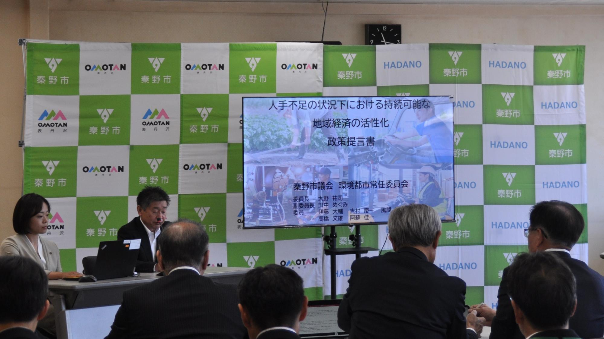 「秦野市」のロゴがある背景の前にスライドが映し出され、テーブルに着席した男女が発表し、スーツ姿の聴衆が聞いている環境都市プレゼンの様子の写真