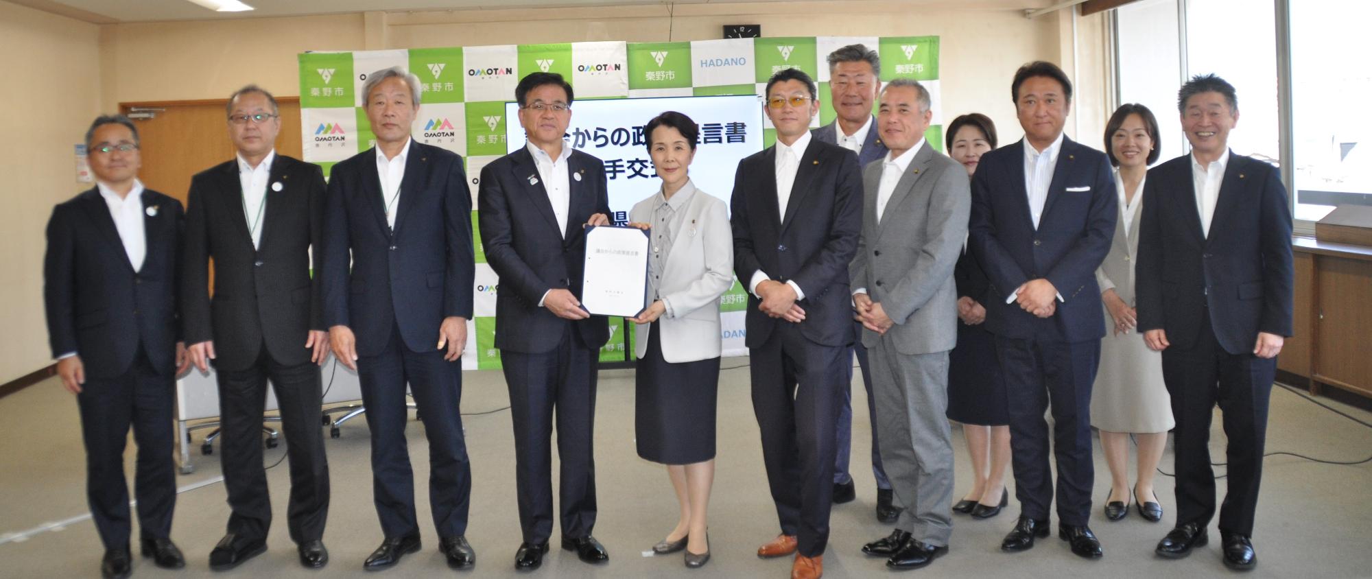 「秦野市」のロゴがある背景の前で、スーツ姿の男女12人が横一列に並び、中央の市長と議長が「議会からの政策提言書」と書かれた書類を一緒に持って微笑んでいる集合写真