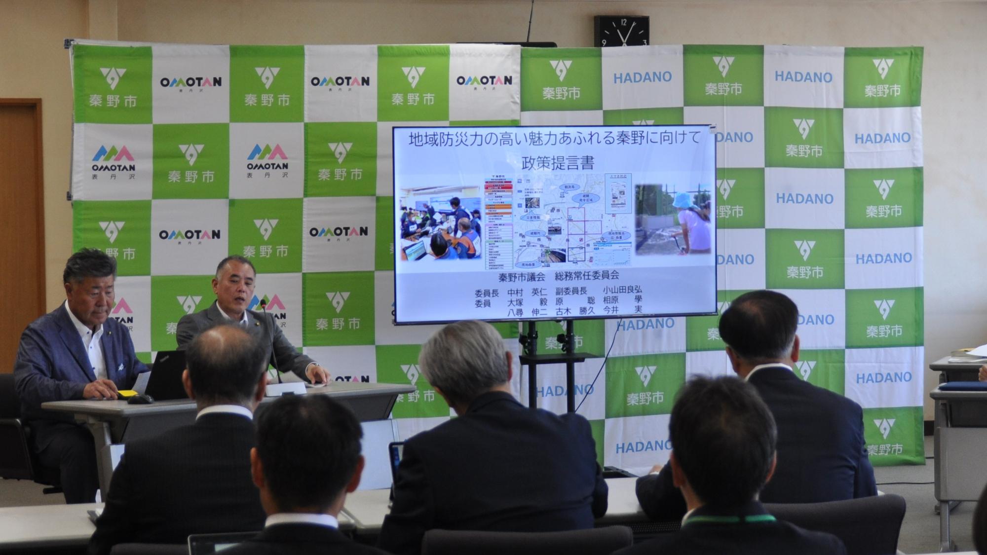 「秦野市」のロゴを背景に、「地域防災力の高い魅力あふれる秦野に向けて 政策提言書」と題したスライドが映る前で男性が発表し、スーツ姿の聴衆が聞いている総務プレゼンの様子の写真