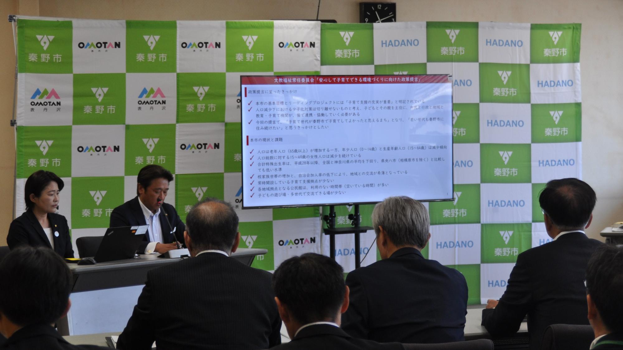 「秦野市」のロゴが並ぶ背景の前にスライドが映し出され、テーブルに着席した男女が発表し、スーツ姿の聴衆が聞いている文教福祉プレゼンの様子の写真外部リンク