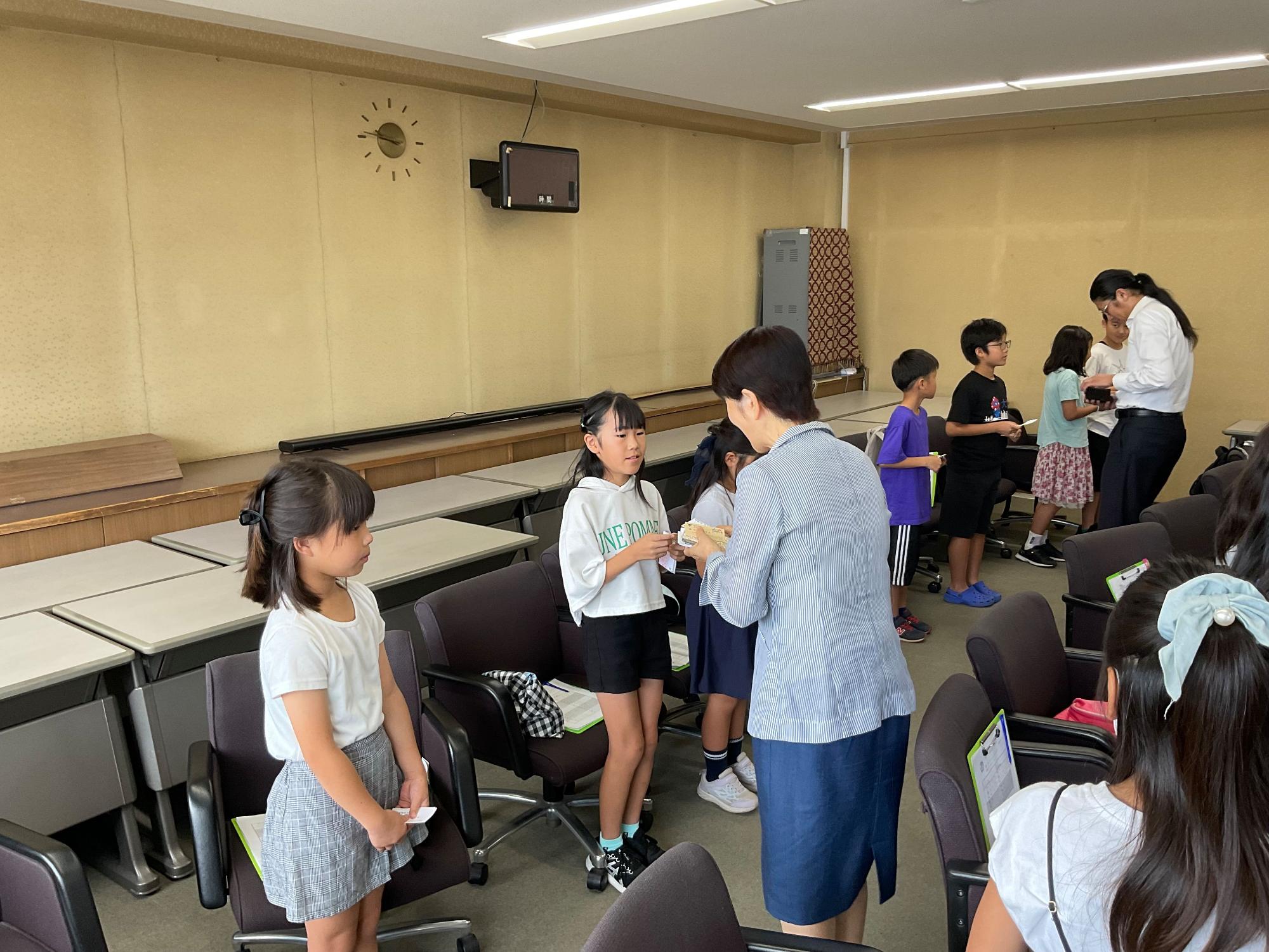広々とした会議室で、女性職員が数人の子供たちに名刺を手渡しており、他の子供たちも列を作って待機したり、スタッフから資料を受け取ったりしている様子を捉えた写真
