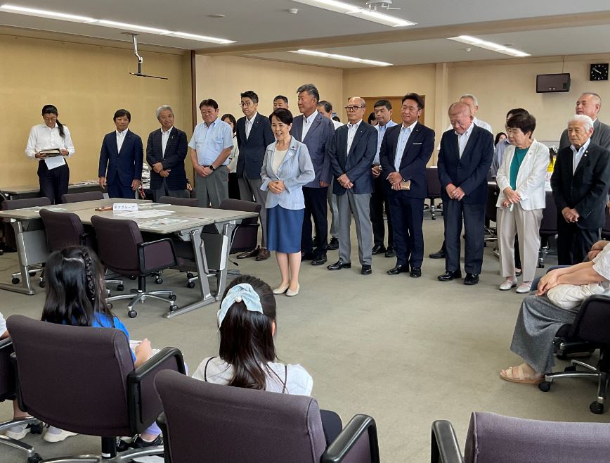 会議室で児童たちが座っている前で、多数のスーツ姿の男性と数名の女性が中央で女性職員が話すのを囲んで立っている写真