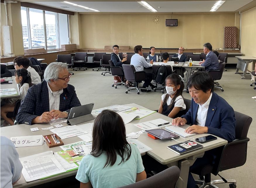 広い会議室で、テーブルごとに分かれた大人と子供たちが、資料やタブレットを使いながら、笑顔でコミュニケーションをとる様子が写っているイベントの写真