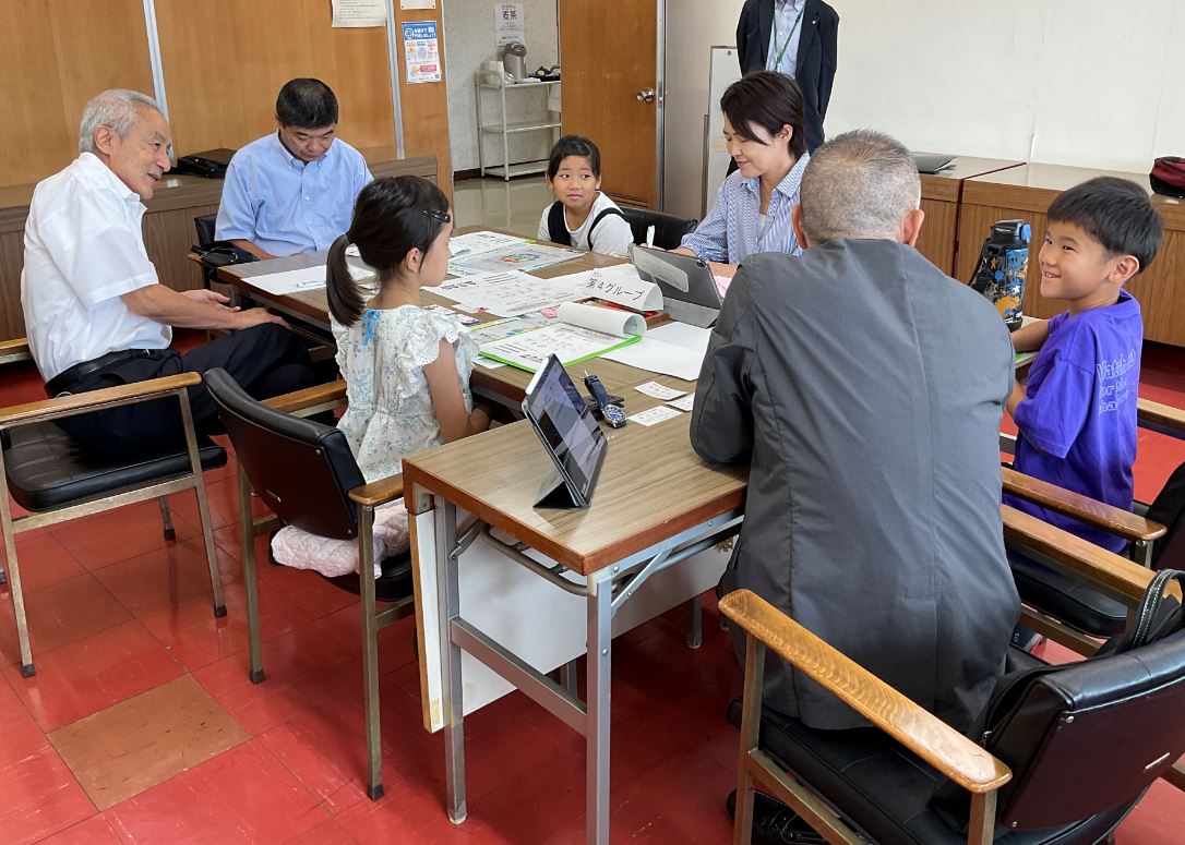 大人と子供たちが会議室のような場所で小さなテーブルを囲み、資料やタブレットを見ながら笑顔で会話を交わしており、世代間交流が行われているイベントの一場面を捉えた写真