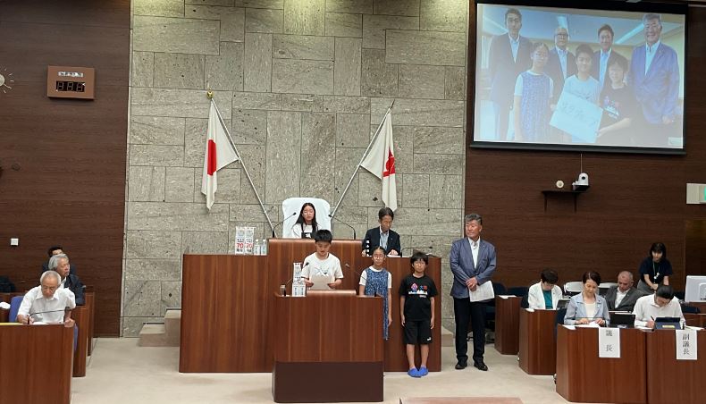 市議会の本会議場のような場所で、中央の演台に3人の児童と、その横にジャケット姿の男性が立ち、スクリーンには児童と大人のグループが写し出されている写真