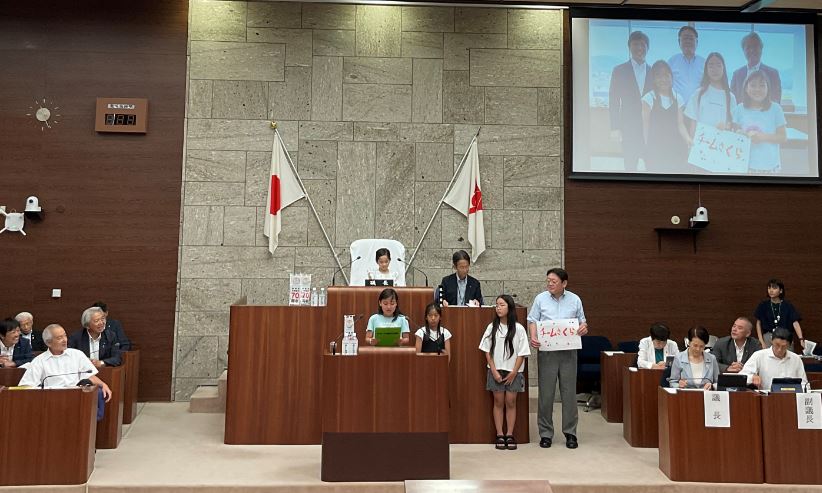 市議会の本会議場のような場所で、中央の演台に3人の児童と、その横に白いボードを持ったの男性が立ち、スクリーンには児童と大人のグループが写し出されている写真