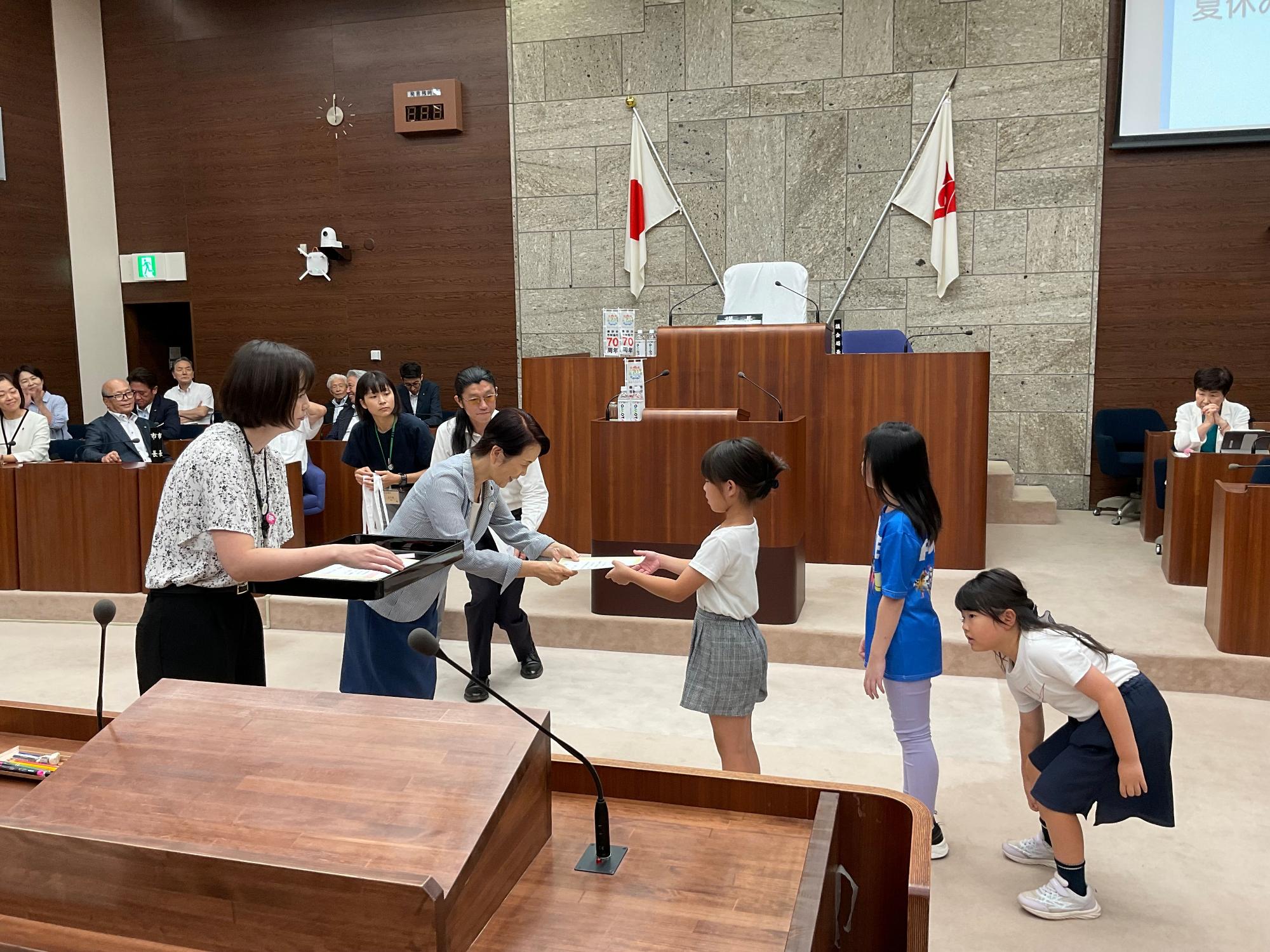 広々とした議場のような空間で、木製の演壇と壇上の席を背景に、二人の女性が大勢の聴衆が見守る中、三人の小学生くらいの女の子たちに終了証を手渡しており、女の子の一人がお辞儀をしている様子を捉えたイベントの写真