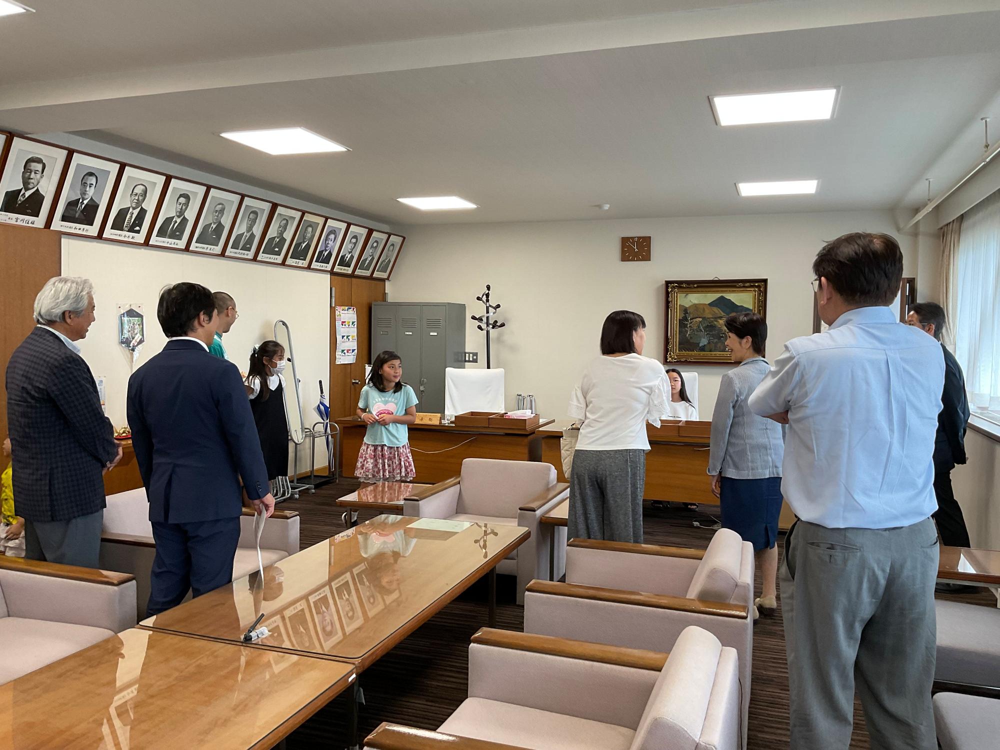 壁には額に入った肖像画が並んだ応接室のような部屋で、複数の大人が立ち、中央に立つ小さな女の子が何かを手に持ち挨拶をしている様子を捉えた写真