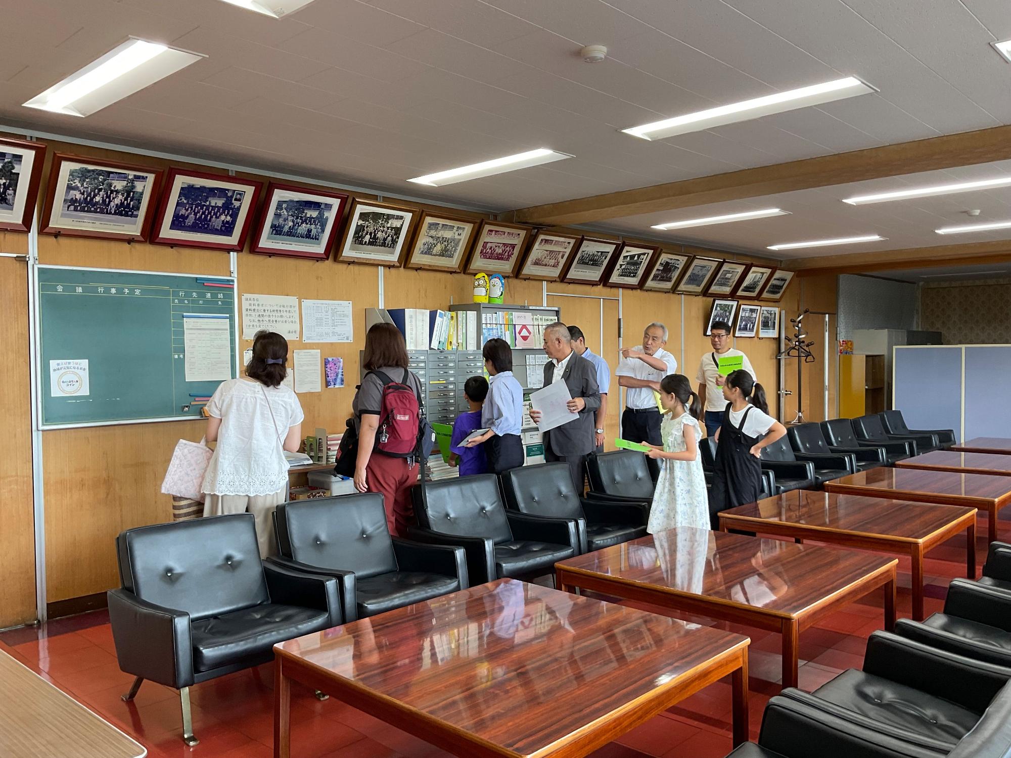 天井照明が明るい公共の待合室のような部屋で、手前に並ぶ光沢のある木製テーブルと黒いソファー席があり、奥の壁際には多くの額装された写真が飾られ、そこに数人の大人と子どもが立ち、書類を見たり話し合ったりしている様子を捉えた室内の写真
