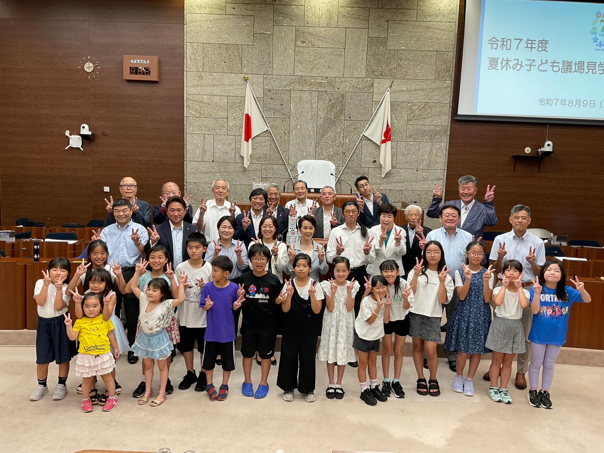 茶色の木目調と石造りの壁が特徴の議場のような場所で、「令和7年度 夏休み子ども議場見学」という文字と日付が映し出されたスクリーンを背景に、大勢の大人と子供たちが前列に並び、全員が笑顔でピースサインをして集合写真を撮影している様子の写真
