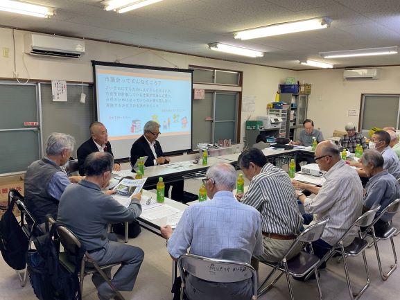 白髪の男性9名ほどがプロジェクターのスクリーンを見ながらテーブルを囲んで座っており、スクリーンには「市議会ってなんだろう」と書かれたプレゼンテーション資料が投影されている様子を捉えた屋内での出前講座の風景写真