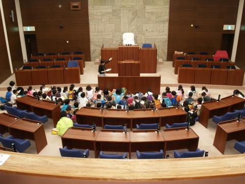 大勢の子供たちが議員席に囲まれた中央の床に座り込み、前方に立つ大人の女性の説明に耳を傾けている議場見学の様子の写真