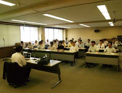 教室のような会議室で多数の人々が机に着席し、前方の講師らしき人物の話を聞いている様子の写真