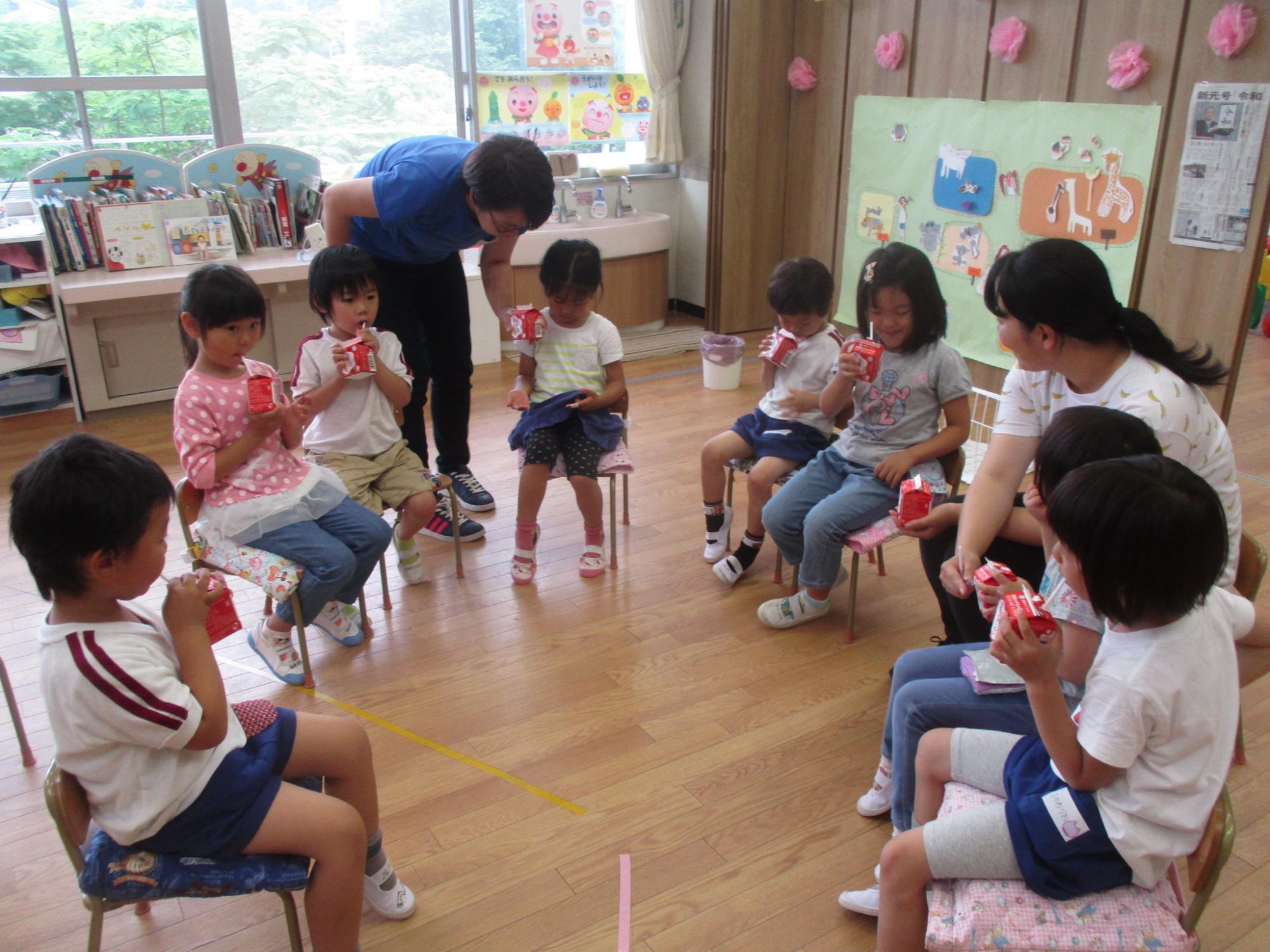 複数の園児が室内で円状に椅子を並べて座り、先生に見守られながら牛乳を飲んでいる写真