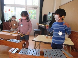 保育室で、マスクを着用した三人の園児たちが、手前の木琴や鉄琴などの楽器を使い、真剣な表情で合奏に取り組んでいる写真