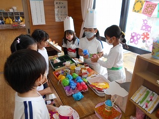 室内のテーブルを囲んで、コック帽をかぶり白いエプロンを着用した3人の園児が、色とりどりの手作りのおもちゃや容器を並べてコックさんになり、向かい側で3人の園児がお客さんになっている様子のごっこ遊びの写真