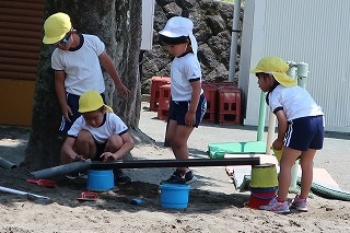 園庭の砂場で体操服と黄色い帽子を着用した複数の園児たちが、バケツを土台にして樋を組み、水遊びをしている様子の写真