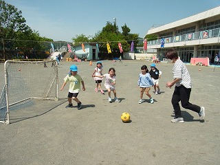 晴れた屋外の園庭で、数人の園児たちが黄色いサッカーボールを追いかけ、サッカーゴールに向かって楽しそうに遊んでいる写真
