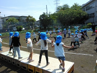 屋外で青い帽子をかぶった5人の園児が前方の台の上で踊り、向かい合わせになって多くの園児たちが楽しそうにダンスを踊っているのを先生も一緒に踊っている様子の写真