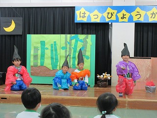 手作りの衣装を着た4人の園児たちが、竹林の背景と「はっぴょうかい」と書かれた看板が飾られた舞台の上で、お遊戯会のような出し物を演じている様子の写真