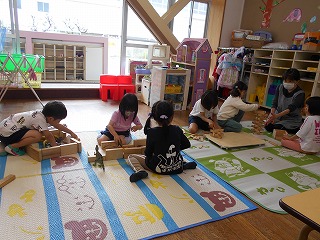 床に敷かれたマットの上に木製の積み木を広げ、6人の幼児が座り込んで夢中になって遊んでいる様子と、その奥で女性保育士が一緒に遊んでいる写真