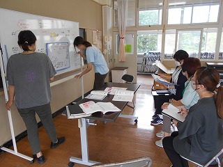 保育者が集まり、二人の女性がホワイトボードに書き込みながら、他の三人が資料やノートを持って着席し真剣な表情でホワイトボードを見ている写真