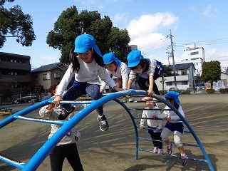 快晴の園庭で、青い帽子を被った園児たちが青い半円型のうんていによじ登って楽しそうに遊んでいるのを横で先生が見守っている写真