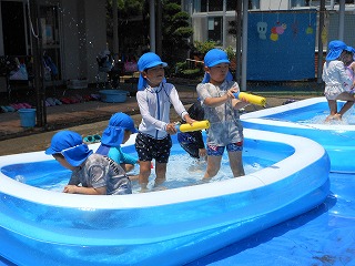 屋外に設置されたビニールプールで、水着を着用し青い帽子を被った複数の園児たちが水鉄砲で遊んだり、プールにしゃがんでいる写真