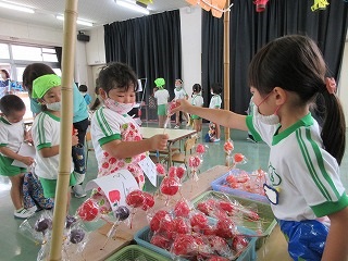 室内で複数の園児たちが夏祭りの屋台で、りんご飴やいちご飴などが置かれ、一人の園児が赤い飴を別の園児に渡している写真
