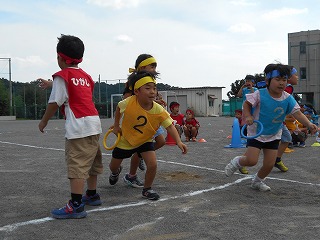 広々とした園庭で、はちまきをつけビブスを着用した園児たちが、丸い園児用のリレーのバトンを持ちリレーをしている写真