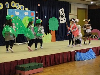 舞台の上で複数の園児たちが「日本一」と書かれた旗や手作りの衣装を身につけ、楽しそうにお遊戯会を披露している写真