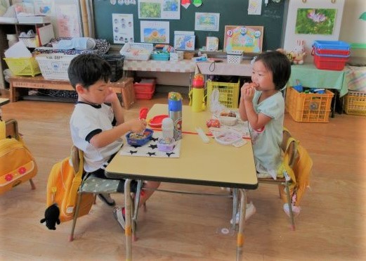 黄色い通園用のリュックが、椅子の背もたれにかかっている椅子に座り、向き合ってお弁当を食べている園児2人を写した写真