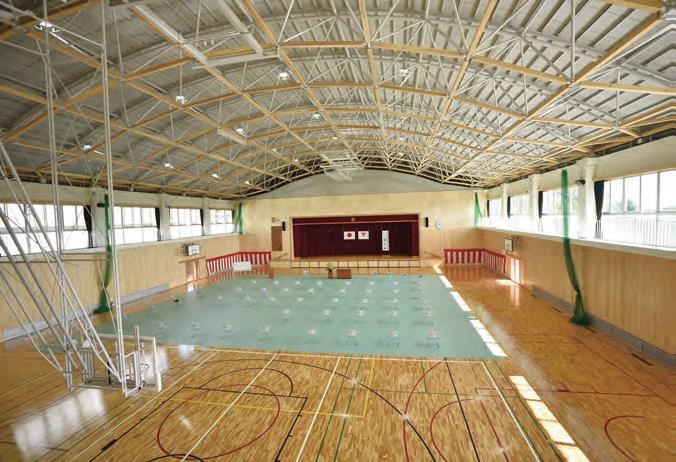 明るい木目の壁と床を持つ広い体育館内部を捉えた写真で、手前にはバスケットコートのラインが引かれ、中央には舞台がある体育館の内部を高い所から全体を写した写真