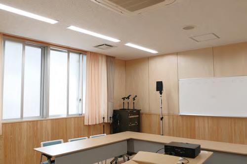 木目調の壁と白い天井の視聴覚室の内部を写した写真で、部屋の奥には窓があり、手前にはU字型に配置された長机と椅子、ホワイトボードなどが映っている写真