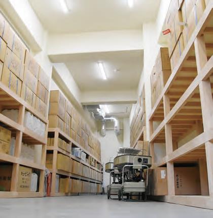 白い壁と天井の部屋に、木製の棚が左右に整然と並び、段ボール箱や備品が隙間なく積まれている防災備蓄倉庫の写真