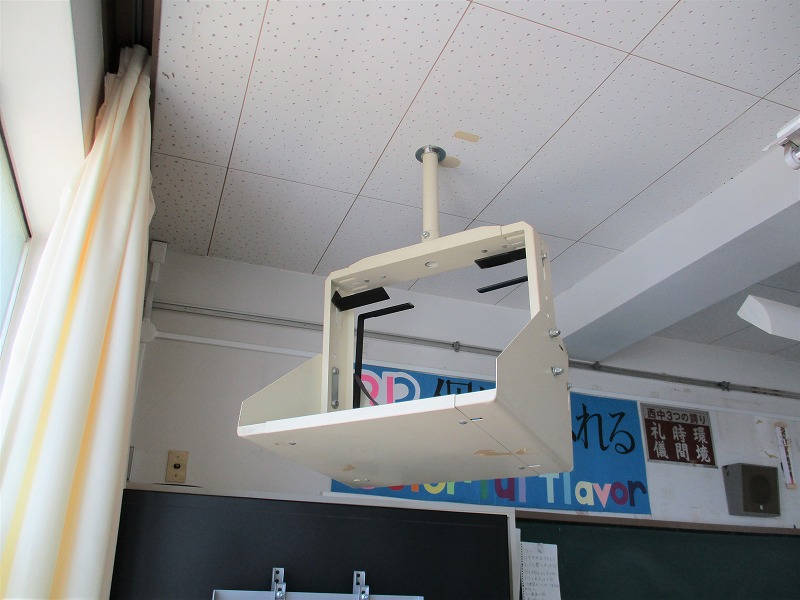 クリーム色のプロジェクター吊り下げ金具が、白い格子状の天井から長い棒で吊り下げられ、左端には黄色の縦縞模様のカーテンと窓枠が見える教室内の写真