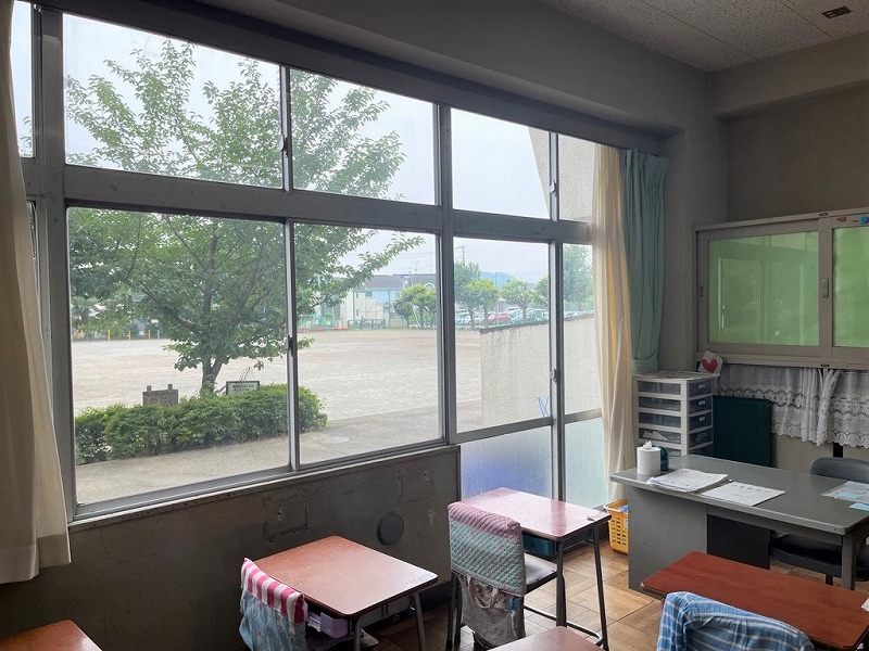 教室の窓辺の様子で、大きな窓の外には木々や建物と車が見える校庭があり、室内には学習机と教卓や書類棚が置かれていて、右側には水色のカーテンが引かれている写真
