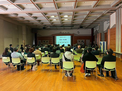 白と黄緑色のスタッキングチェアが整然と並べられた広い室内で、多数の参加者が着席してステージのスクリーンに映し出された資料を聴講している講演会の様子の写真