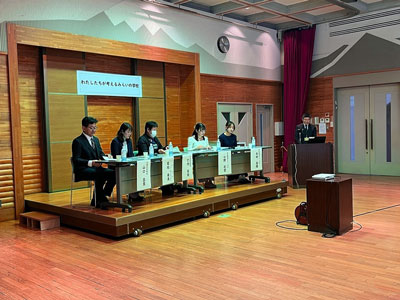 小上がりの舞台に着席している5人のパネリストと右側には演台の前にマイクスタンドの司会者が立ちディスカッションしている様子の写真