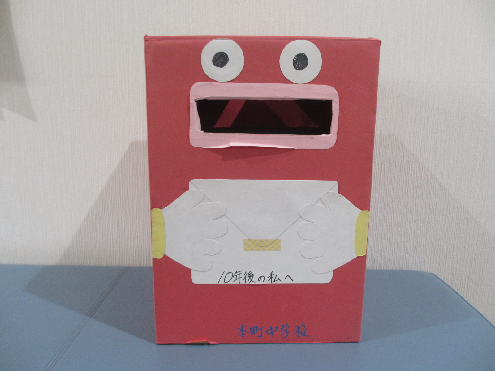 白い背景の前で青い台の上に置かれた、赤い段ボール箱で作られた顔のポストで、白い円の目とピンクの口、白い手袋をした両手に「10年後の私へ」と書かれた封筒を持ち、底部に「本町中学校」と記された工作作品の写真