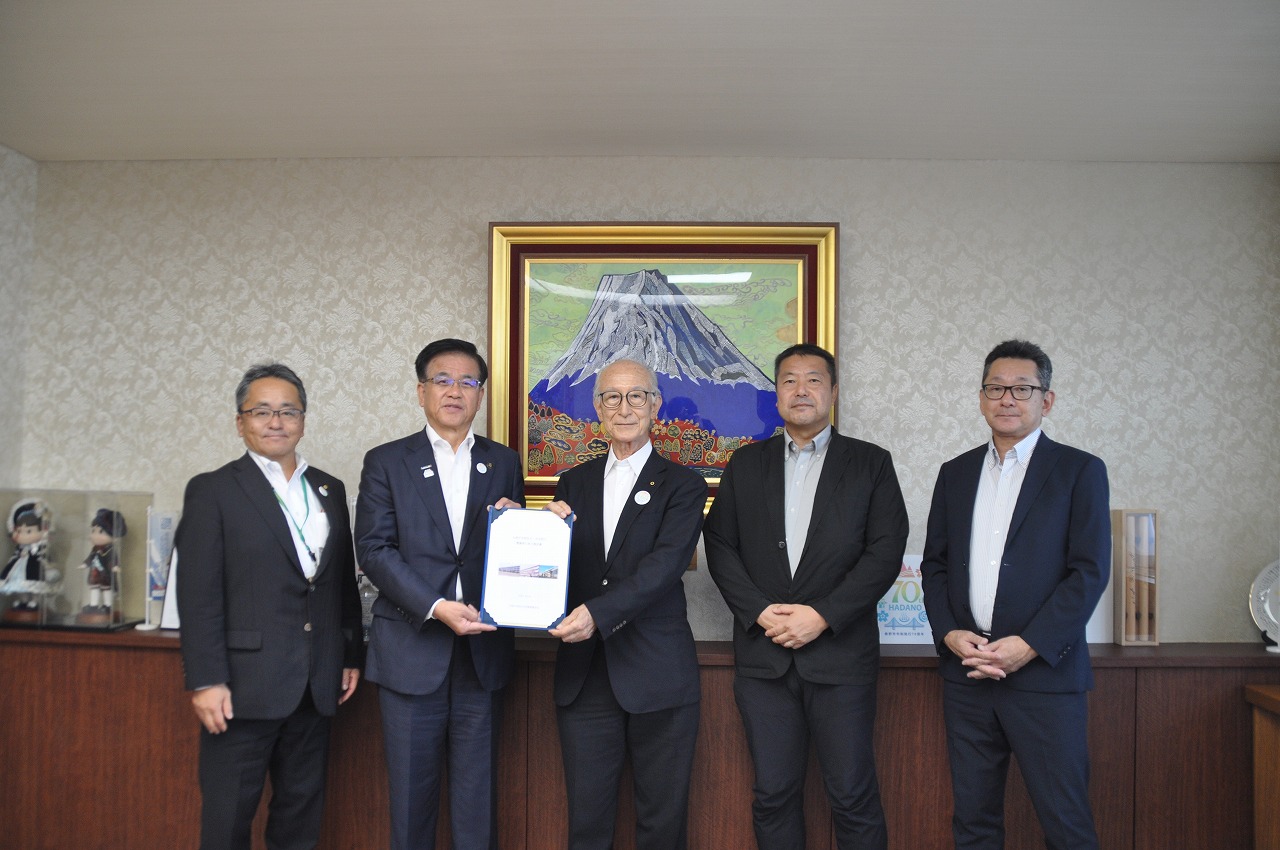 富士山の絵画を背にした室内の木製カウンター前で、スーツやジャケット姿の5人の男性が並び、中央の市長と男性が証明書のような書類を一緒に持ってカメラに向かって立っている集合写真