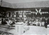屋内の広間に着物姿や洋服姿の人々が大勢集まり、床に座り込んだり立ったりしており、奥には旗が掲げられている様子が確認できる、粒子の粗い白黒の古い写真