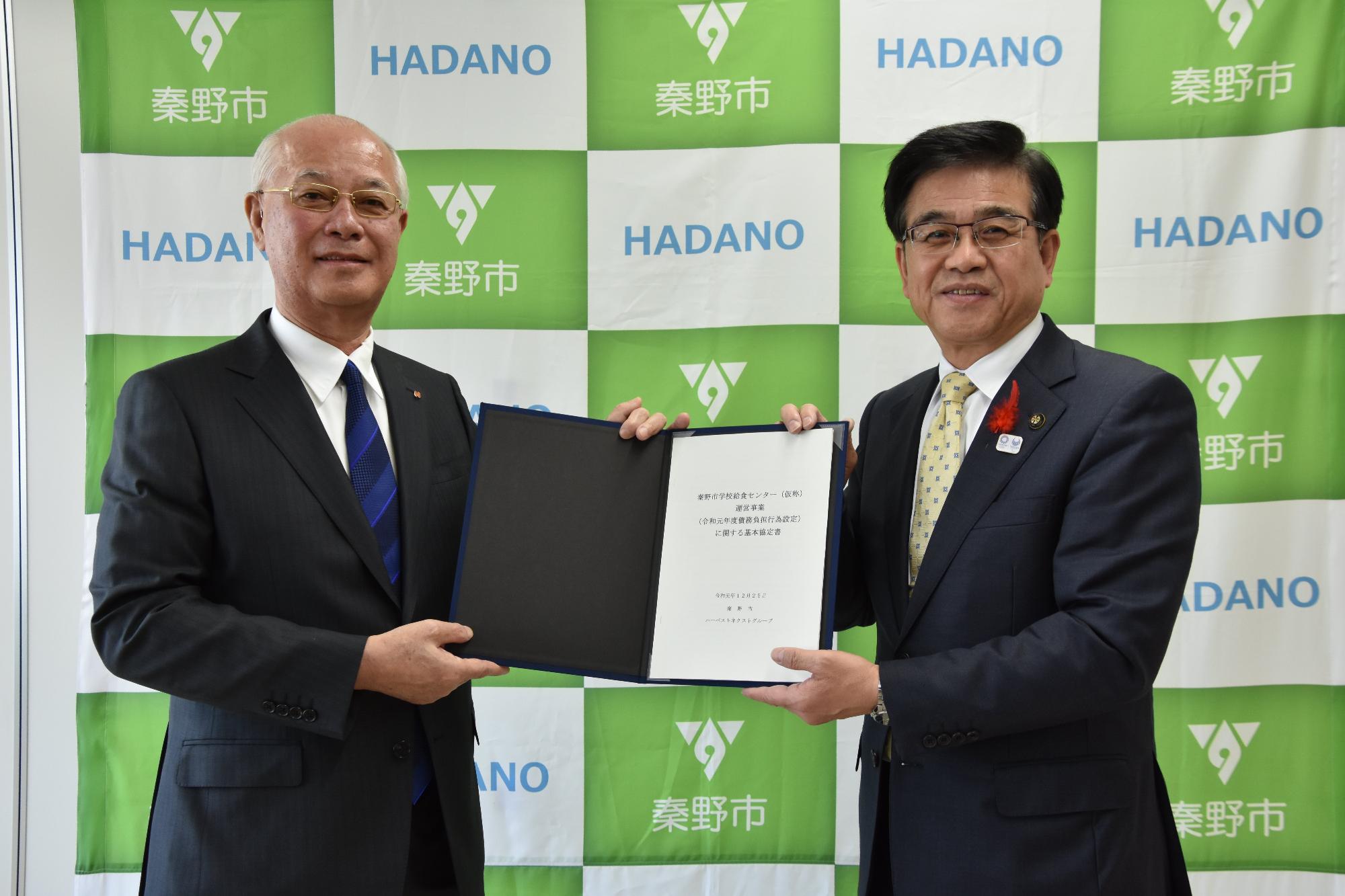 ネイビーのスーツを着た市長と男性が、秦野市のロゴが入った緑と白の背景の前で、黒いファイルに入った文書を両手で持ちながらカメラに向かって微笑んでいる写真