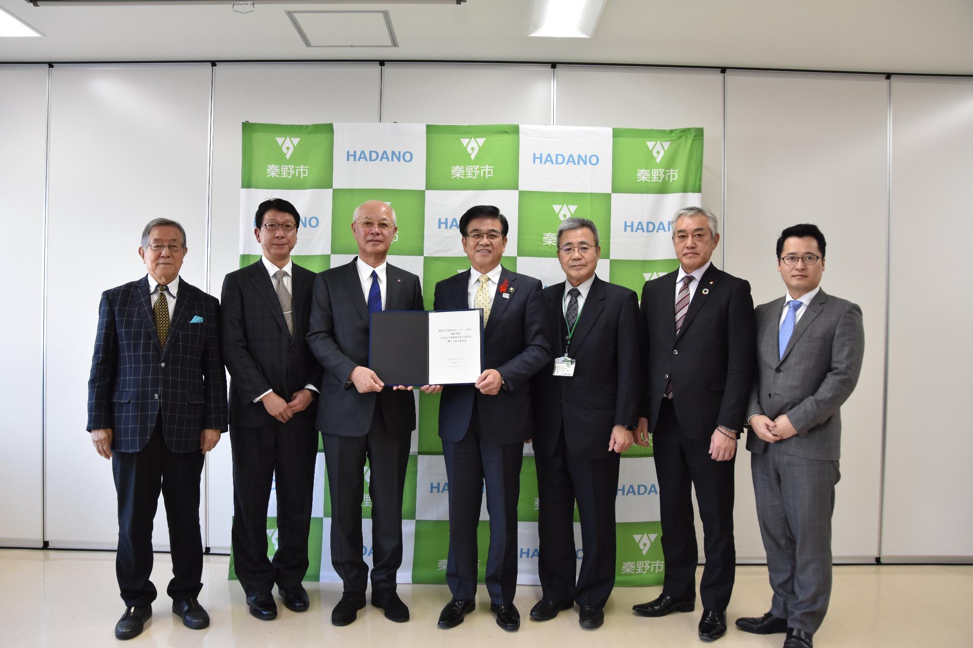 秦野市のロゴが入った緑と白のパネルの前で、中央の2人が黒いファイルに入った文書を持ち、両側にそれぞれ二人の男性が立ち、全員がスーツ姿でカメラに向かって立つ集合写真