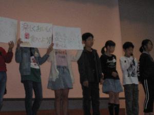 ステージ上で、子供たちが「楽しくおいしく食べよう」などと書かれた白いプラカードを掲げたり、マイクを使って発表をしている様子の写真
