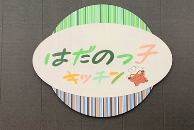 緑やオレンジなどの優しい色合いの文字で「はだのっこキッチン」と書かれ、右下にボンチーヌが描かれた楕円形の看板の写真